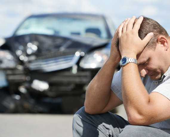 crash A man with his head down holding his head with a crashed car in the background